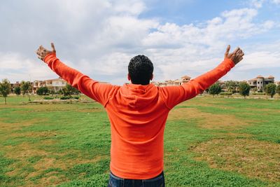 Rear view of man standing with arms outstretched on field against cloudy sky