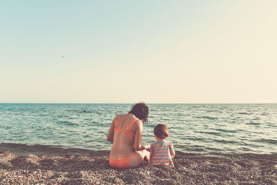 Rear view of mother sitting with daughter on shore at beach