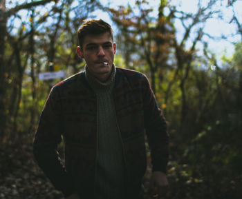 Portrait of young man smoking cigarette while standing against trees