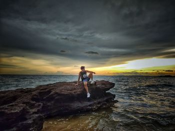 Man on rock by sea against sky during sunset