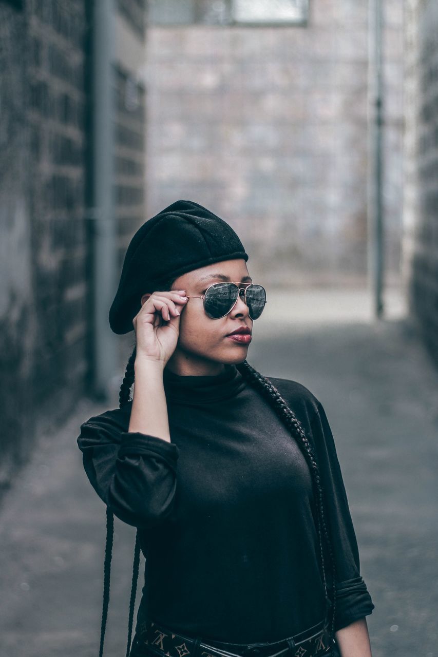 YOUNG WOMAN WEARING SUNGLASSES STANDING AGAINST WALL