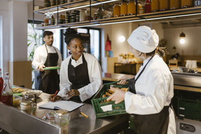 Female chef holding crate and taking inventory with colleague in commercial kitchen at restaurant