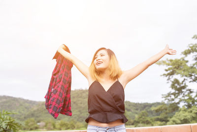 Young woman with arms raised standing on field against sky