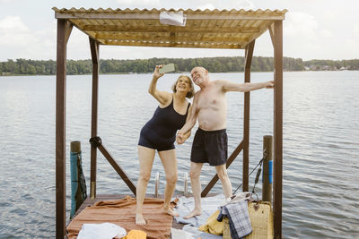 Full length of senior woman taking selfie with man while standing in gazebo against river