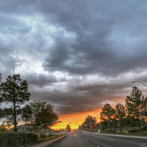 Road by trees against dramatic sky during sunset