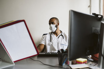 Male doctor wearing protective face mask reading medical files while sitting at desk