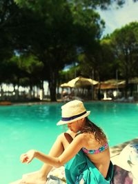 Side view of girl wearing hat while sitting at poolside against trees