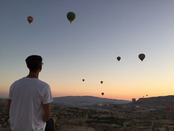 Rear view of man looking at hot air balloons flying over landscape during sunset