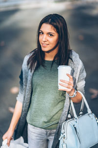 Portrait of young woman holding disposable cup while standing in city
