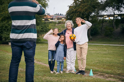 Midsection of man photographing happy grandmother and grandchildren gesturing peace sign in park during picnic