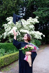 Happy woman in graduation gown throwing mortarboard while standing against plants