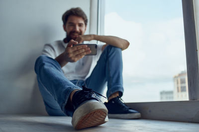 Young man using mobile phone while sitting on window