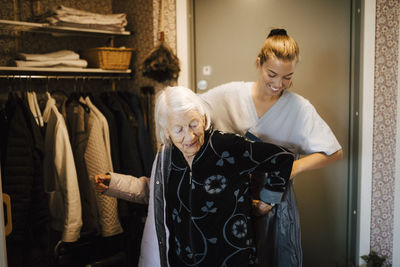 Smiling female nurse heaping senior woman to wear jacket at home