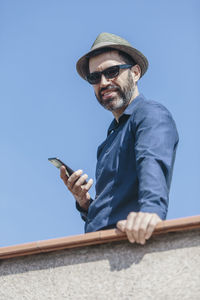 Attractive middle-aged man with hat and sunglasses talking to a smart phone standing on balcony.