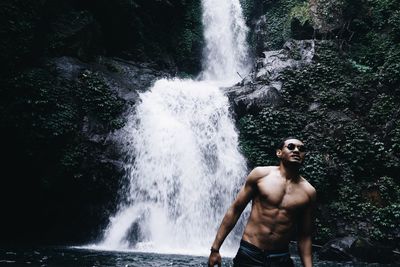 Shirtless man standing against rock and waterfall