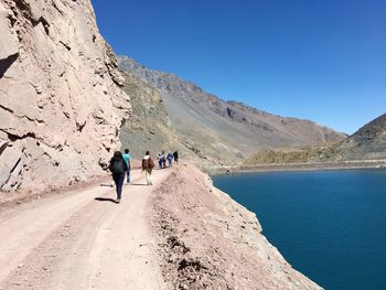 People walking on dirt road by lake against clear blue sky