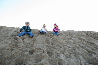 Siblings sitting on sand dune at beach against sky