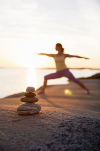 Woman practicing yoga warrior pose on lakeshore with focus on stack of stones