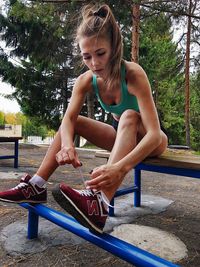 Young woman tying shoelace while sitting on bench against trees in park