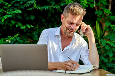 Happy mature male manager in white shirt speaking on smartphone and writing notes in planner while sitting at table with laptop during work in street restaurant in summer