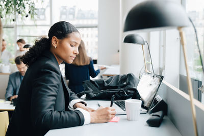 Businesswoman writing notes while working on laptop at desk in coworking space