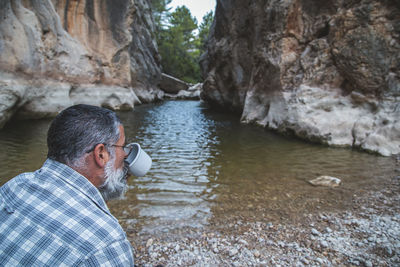 A bearded tourist drinking from a travel mug sitting on the river bank on a steep background