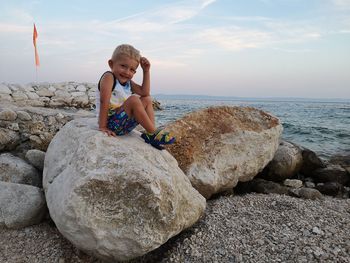 Portrait of smiling cute boy sitting on rock against sea during sunset