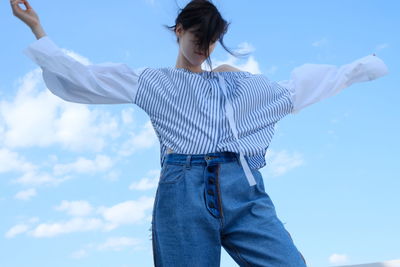 Low angle view of young woman with arms outstretched standing against sky