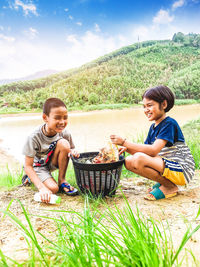 Smiling siblings with basket crouching on land by lake