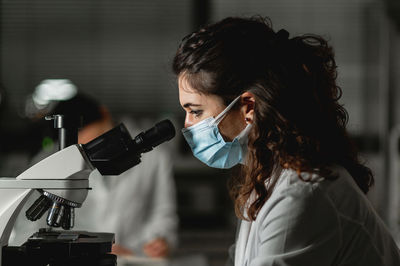 Side view of skilled female scientist in protective mask using microscope while conducting chemical experiment in modern lab