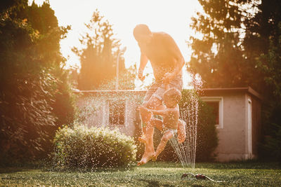 Father and child jumping over the water from the sprinkler in garden