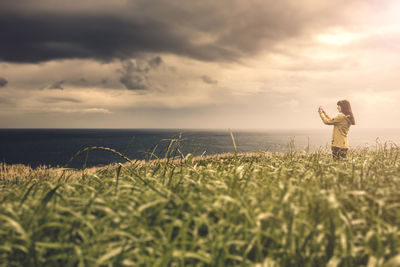 Woman photographing sea while standing amidst plants during sunset