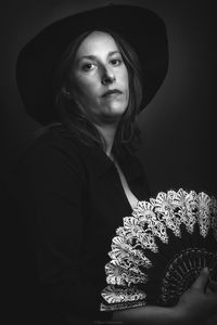 Portrait of woman wearing hat holding folding fan standing against black background
