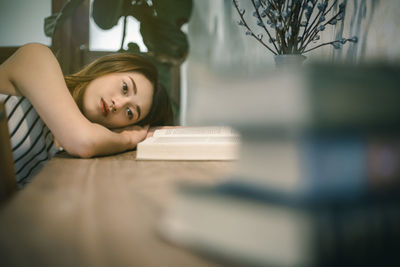 Thoughtful young woman lying by book on table