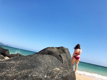 Rear view of woman in bikini standing on shore at beach against clear blue sky