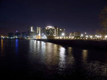 Illuminated buildings by river against sky at night