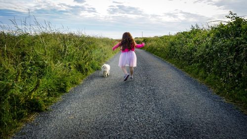 Rear view of girl and dog running on road amidst plants against sky