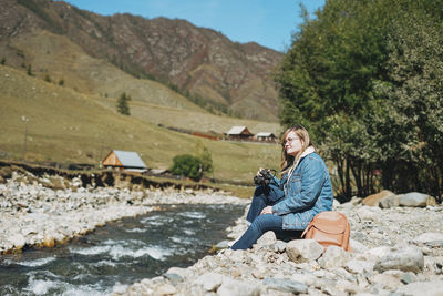 Beautiful smiling blonde young woman traveler with photo camera in hands near mountain river