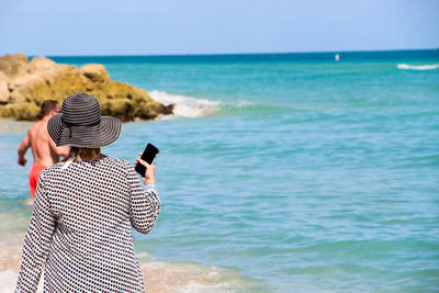 Rear view of woman holding mobile phone at beach on sunny day