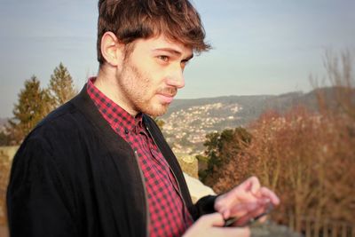 Close-up of thoughtful young man using phone while standing against sky