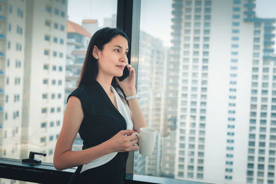 Businesswoman holding coffee cup and using phone while standing by window