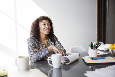 Smiling businesswoman sitting by table and looking away in office