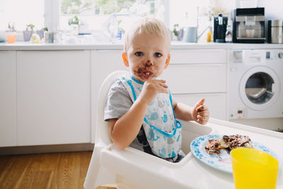 Adorable toddler with face covered in chocolate sitting on table at home
