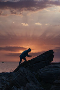 Man on rock by sea against sky during sunset