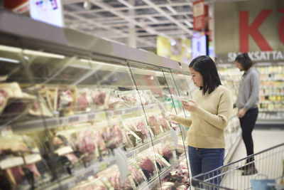 Woman doing shopping in supermarket and using cell phone to compare prices or checking shopping list