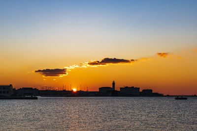 Silhouette buildings by sea against sky during sunset