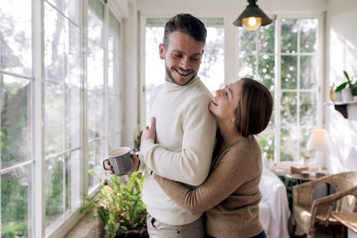 Side view of cheerful female embracing bearded male beloved with mug of coffee while looking away against window in house