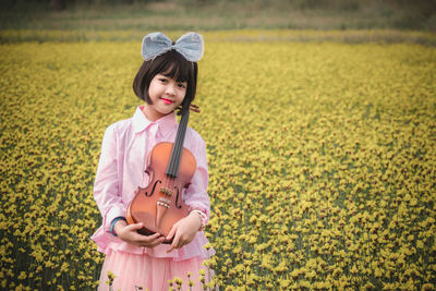 Portrait of smiling cute girl holding violin while standing amidst flowering plants on field