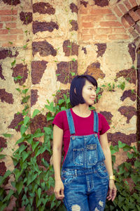 Smiling young woman looking away while standing against ivy on wall