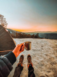 Low section of person holding coffee mug against sky during sunset
